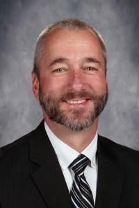 Smiling man with short gray hair and beard wearing a black suit, white shirt, and striped tie in front of a gray studio background.